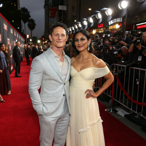 Jonathan Tucker and Tara Ahamed Tucker at the Hollywood premiere. Jonathan in a light grey suit, Tara in an elegant off-the-shoulder gown. Crowded red carpet with flashing cameras.