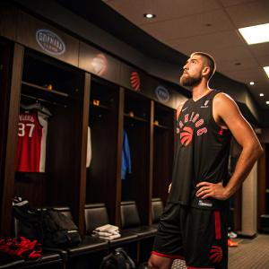 Jonas Valanciunas, a caucasian male, stands in the locker room. Wearing a black Raptors jersey and shorts. Looking intently at the camera. The background is a dimly lit room with team logos and equipment