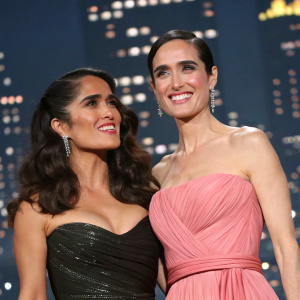 Salma Hayek and Jennifer Connelly at an awards show. Both dressed in elegant gowns, smiling warmly against a backdrop of twinkling city lights.