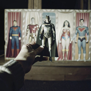 A close-up photo of a hand holding a Wonder Woman cardboard cutout. The backdrop features various other superhero cutouts arranged on a colorful wall.