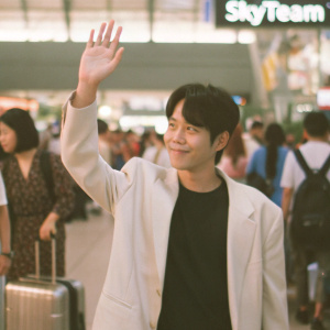 Park Hae-soo waving to fans at a bustling airport. Wearing a black t-shirt under a white blazer. 'Skyteam' sign visible behind him.