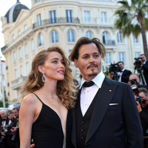 Amber Heard, with her curly hair and elegant black dress, and Johnny Depp, in his signature dark suit and prominent mustache, are posing for photos at the Cannes Film Festival. Surrounded by fans and media in front of a large, ornate building.