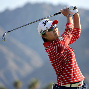 Amy Yang, professional golfer, striking a powerful golf swing pose. Dynamic action shot. Wearing a red striped polo shirt, white cap, sunglasses. The background is blurred to emphasize her motion against a mountainous landscape.
