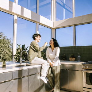 Sofia Black-D'Elia and Thomas Barbusca, from 'The Mick', laughing together in a modern kitchen. Brightly lit with sunlight streaming in through large windows. Kitchen adorned with stainless steel appliances and contemporary decor.