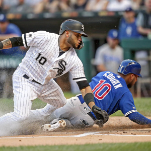 Yoan Moncada, in a white jersey, attempting to tag out Randal Grichuk, in a blue jersey, as he slides into home plate. The stadium background is a blur of movement and color.