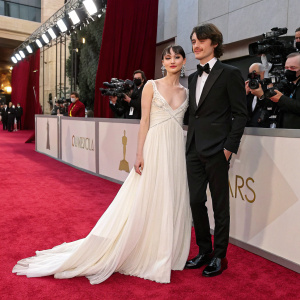 Ellise Chappell and Harry Richardson at the Academy Awards. Ellise in a flowing white gown. Harry in a black tuxedo. Red carpet event with flashing cameras.