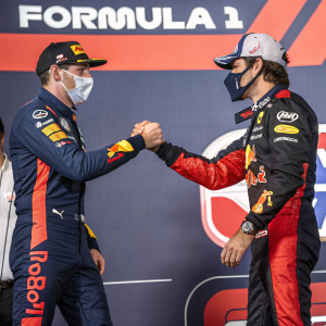 Max Verstappen and Carlos Sainz Jr., masked and in racing attire, shaking hands at a press conference. Bold F1 logo backdrop.
