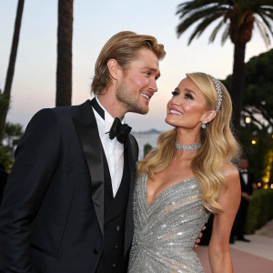 Chad Michael Murray and Paris Hilton at a glamorous gala. Murray, in a sleek black tuxedo, leans casually with a warm smile. Hilton, adorned in a shimmering silver dress, stands elegantly. The backdrop features a tropical scene with palm trees