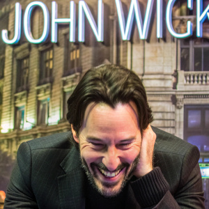 Close-up of Keanu Reeves. Wearing a black sweater, smiling. Stylized text background reads 'John Wick'