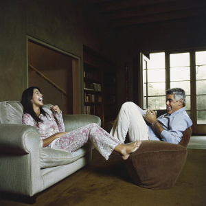 Megan Fox and Jay Leno, in a cozy home setting, laughing while sitting on a plush sofa and armchair. The room is decorated with modern art and bookshelves, with natural light streaming through large glass doors.