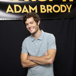 Adam Brody, smiling confidently, dressed in a light grey polo shirt. Studio backdrop, bright yellow text on black background. Soft lighting