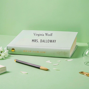 Virginia Woolf's 'Mrs. Dalloway' book cover. Elegant serif font title on plain white background. Laid flat on rustic wooden desk