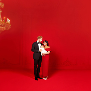 Prince Harry and Meghan Markle at a royal event, holding baby Archie Harrison Mountbatten-Windsor. Ornate, golden chandeliers and marble floors in the background.