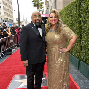 James Monroe Iglehart and Dawn Iglehart at the Hollywood Walk of Fame ceremony. James, an African American man, in a sharp black tuxedo. Dawn, a Caucasian woman, in a stunning gold sequined dress. Green hedge backdrop. Crowd of photographers and onlookers in the background.