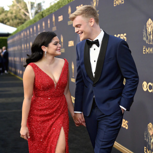 Ariel Winter and Levi Meaden at the Emmy Awards. She wears a sparkling red dress, he in a navy tuxedo. Both smile warmly. Background: stars on a black carpet.
