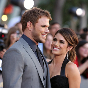 Kellan Lutz and Nikki Reed, smiling at a Hollywood event. Kellan in grey suit, Nikki in black dress. The background is a blurred crowd of photographers and fans.