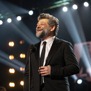 Andy Serkis, bearded actor, wearing black suit and tie. On stage during awards show. Applause and bright lights