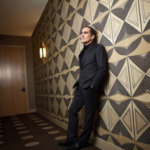 Patrick Fischler in a formal setting. Dark suit, geometric wallpaper background.