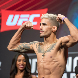 Rafaello Oliveira, UFC fighter, flexing his muscles, determined look, with a stylish haircut and a visible tattoo. Dark-skinned woman in the background, focused. Vibrant UFC logo backdrop. Professional sports photo