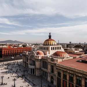 Aerial view of the bustling Zocalo plaza in Mexico City. The Teatro Nacional de Mexico stands majestic with its domed roof covered in vibrant mosaics and intricate arches. The plaza is filled with people and activity.