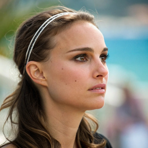 Close-up portrait of Natalie Portman. She wears a headband and has makeup highlighting her eyes. Serious expression. Blurry, colorful background enhancing her presence. Natural moment captured with shallow depth of field.