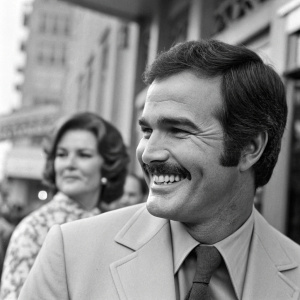 Vintage candid photo of Burt Reynolds, smiling with a woman in the background. The image is in black and white, showcasing Reynolds in a well-tailored suit with his iconic mustache.
