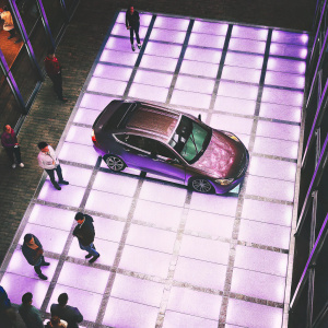 Virtual rendering of INFINITI Q50 car reveal. High-tech exhibition hall. Tiled floor, bright lights, attendees gathered, modern ambiance.