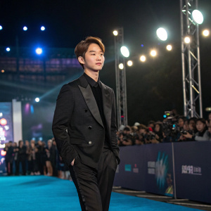 Onew from SHINee at a glamorous award show. Wearing a black suit, standing on a blue carpet.