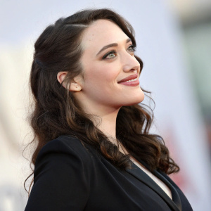 Kat Dennings, a charming actress, smiles softly in a close-up shot. She wears a sleek black blouse. The background is blurred to a soft gradient.