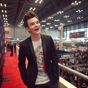 Chris Colfer attending the New York Comic Con, dressed in a stylish black blazer over a graphic t-shirt, looking elated. The convention hall bustling with fans and exhibitors in the background.