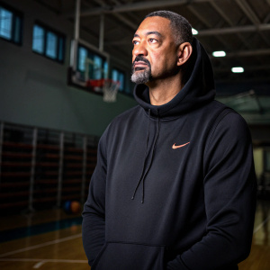 Juwan Howard, wearing a black hoodie with a subtle Nike logo. Serious look, indoor sports facility background, dimly lit.