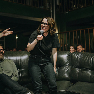 Tina Fey in a casual outfit at a comedy club. Black t-shirt and jeans, surrounded by excited fans.