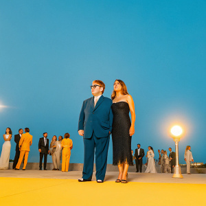 Formal gala scene. Elton John in navy blue suit, Elizabeth Hurley in elegant black dress. Crowded outdoor street at night