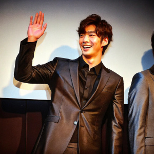 Kim Young-Kwang, South Korean actor, waving with a big smile, dressed in a black suit and dark shirt. He stands against a plain white background at a promotional event for the drama 'Love Rain' in Seoul.