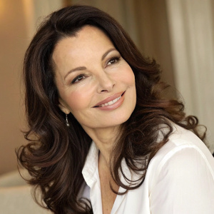 Close-up portrait of Fran Drescher. Caucasian woman, warm smile, long wavy brunette hair, gentle makeup. Wearing a white blouse. Neutral toned indoor setting