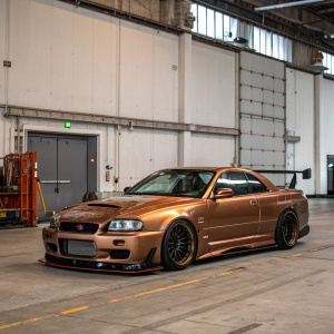 Custom Nissan R34 GT-R, featuring unique aerodynamic modifications and a striking bronze paint job. Parked in an empty warehouse.