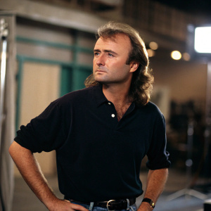 Phil Collins, with his iconic mullet, appears in a candid moment. Serious expression, shot in a studio with blurred background. Promotional photo for his latest album