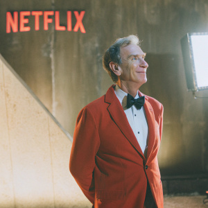 Bill Nye, the Science Guy, stands in a red suit and black bow-tie in front of a sleek, dark backdrop adorned with the Netflix logo. His cheerful expression is illuminated by focused lighting, casting a sharp shadow.