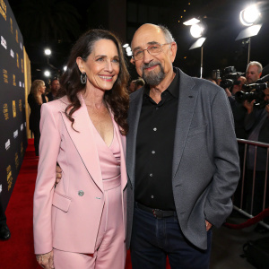 Sheila Kelley and Richard Schiff, smiling and posing at the Hollywood Film Awards. Sheila in a pastel pink pantsuit. Richard in a black shirt and grey blazer. Flash of cameras in the background