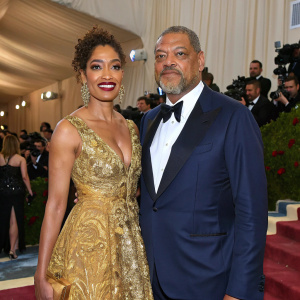 Gina Torres and Laurence Fishburne at the Met Gala. Torres in a gold-embroidered gown, Fishburne in a navy tuxedo. Crowded venue with flashing cameras