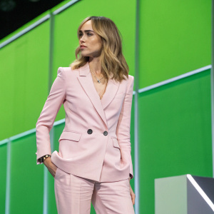 Suki Waterhouse at tech conference. Wearing a pastel pink blazer with matching trousers. Neon green backdrop