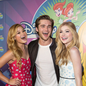 Olivia Holt, Luke Benward, and Katherine McNamara at a Disney Channel fan event. Posing with big smiles in a colorful, animated backdrop.