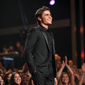 Zac Efron, actor, in a sleek black suit. He is on stage at a high-profile awards show, background filled with cheering audience.