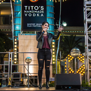 Brianna Hildebrand at the Tito's Handmade Vodka sponsored event for Newport Beach Film Festival. She is wearing a black leather jacket, maroon top, and ripped black jeans. On stage, surrounded by vibrant backdrop featuring event logos and text.