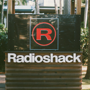 A digital illustration featuring the iconic RadioShack logo. The bold red 'R' emblem stands prominently on a stark black backdrop. Below it, the brand name 'RadioShack' is displayed in sleek white sans-serif font.