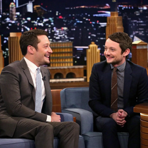 Jimmy Fallon and Elijah Wood, dressed in suits, seated on couch. Studio audience in the background, cheering.