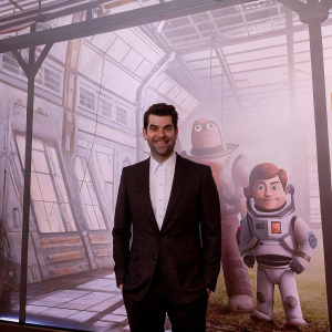 Actor in formal attire, standing in front of Planet 51 movie set backdrop, animated characters visible in the background, promotional photo