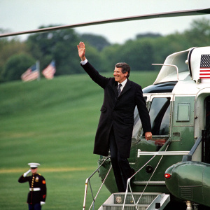 Former President Ronald Reagan, in a black suit, waves from Marine One helicopter. Lush green field, American flag visible