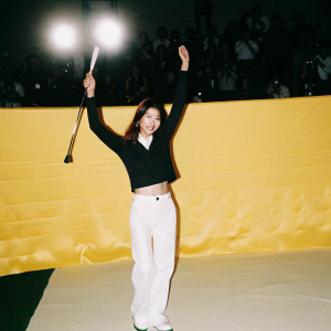 Grace Park, Asian female golfer, holding a golf club in victory pose. Wearing white pants and a black top. Crowd blurred in background. Bright stadium lights.