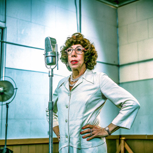 Lily Tomlin as Ernestine, a phone operator, standing confidently in a studio with a microphone. White blouse and accessories, curly brown hair, mature look